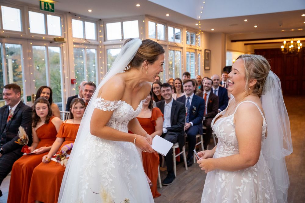 Two brides share a joyful laugh during their wedding ceremony surrounded by guests.