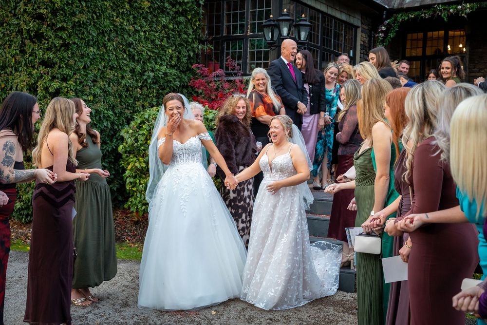 Two brides in white gowns hold hands, surrounded by bridesmaids in green and burgundy dresses at an outdoor wedding.
