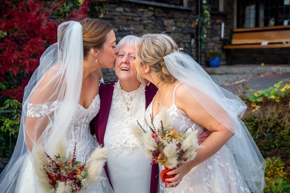 Two brides in white gowns and veils kiss an elderly woman in burgundy at an outdoor autumn wedding celebration.