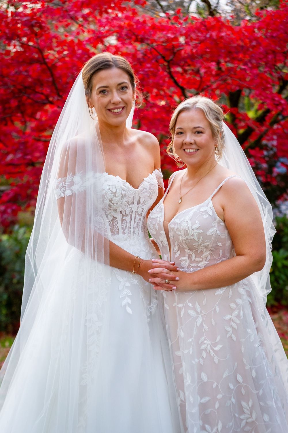Two brides in white lace wedding gowns pose together before vibrant red autumn foliage.
