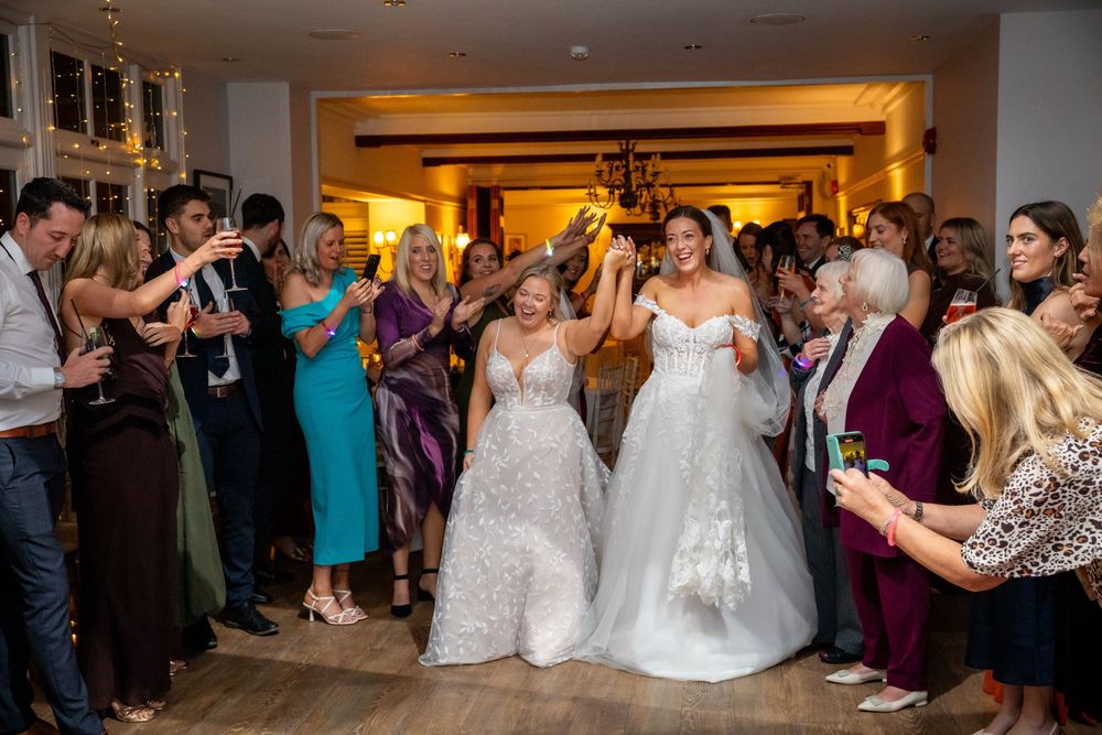 Two brides in white gowns celebrate their wedding reception entrance surrounded by cheering guests.