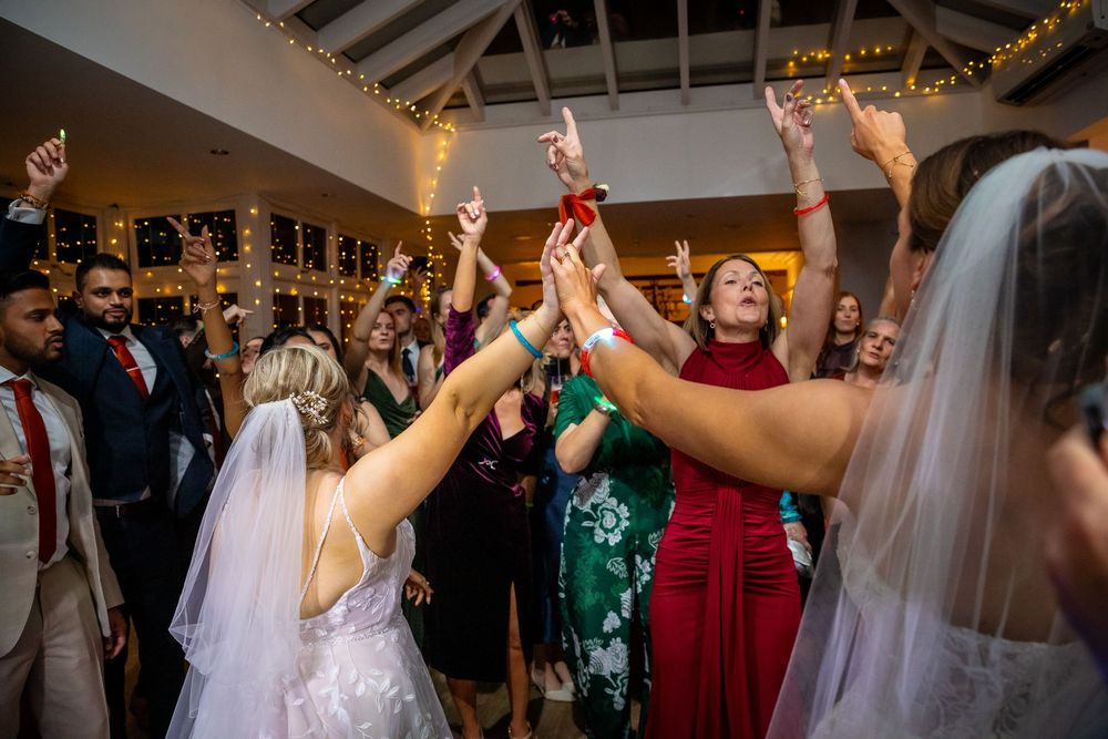 Wedding guests and brides celebrate with hands raised on the dance floor at reception.