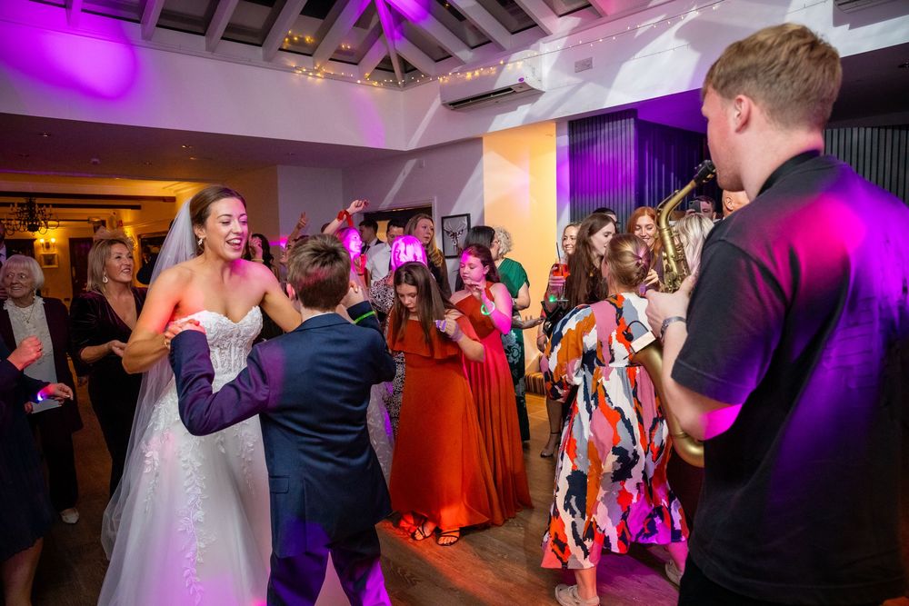 Bride dances joyfully with wedding guests under purple lights at lively evening reception.