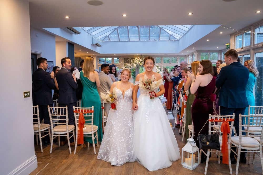 Two brides in white gowns walk down the aisle together at their wedding ceremony surrounded by cheering guests.