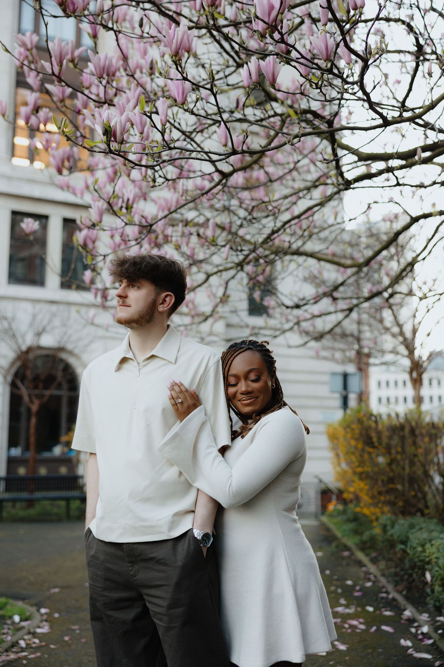 Two people in white outfits stand together under blooming pink magnolia trees on a spring day.
