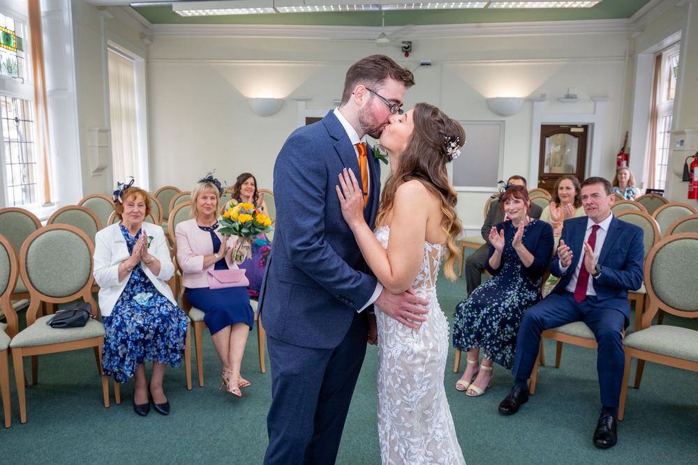 Newlyweds share first kiss as guests applaud in a bright registry office ceremony room.