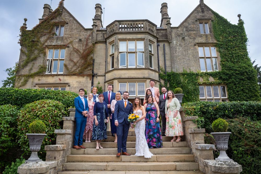 Wedding party posing on stone steps outside a grand manor house with lush gardens.