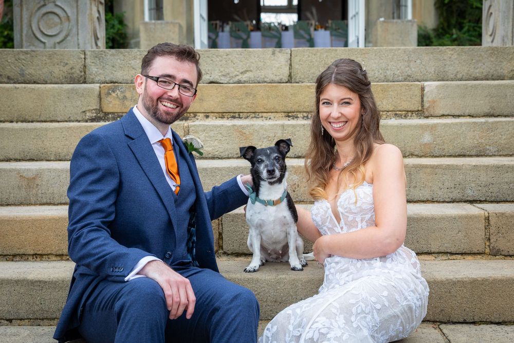 Smiling bride and groom sitting on stone steps with their pet dog at wedding venue.