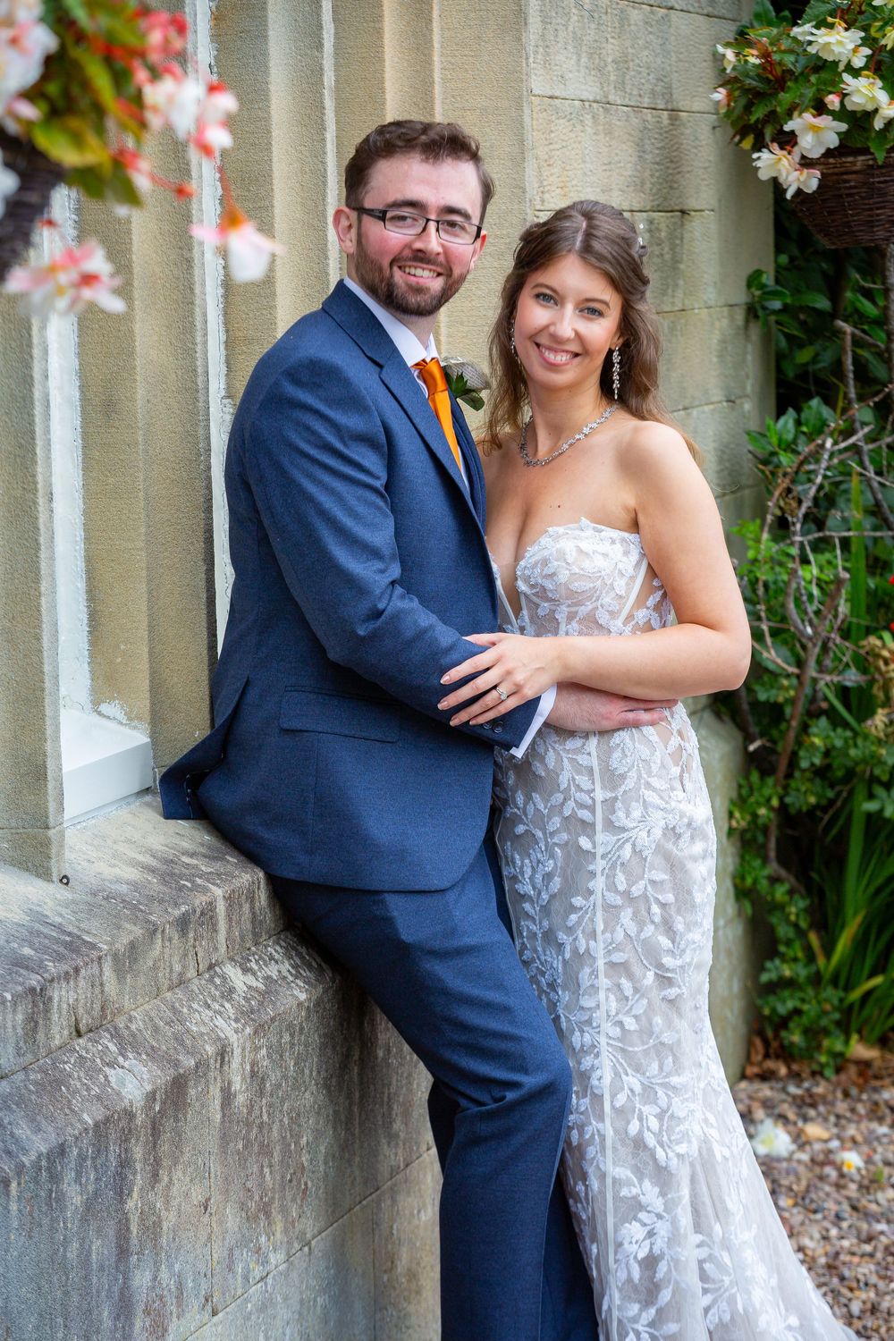 Smiling bride and groom pose outdoors against stone wall with pink flowers, groom in navy suit with orange tie.