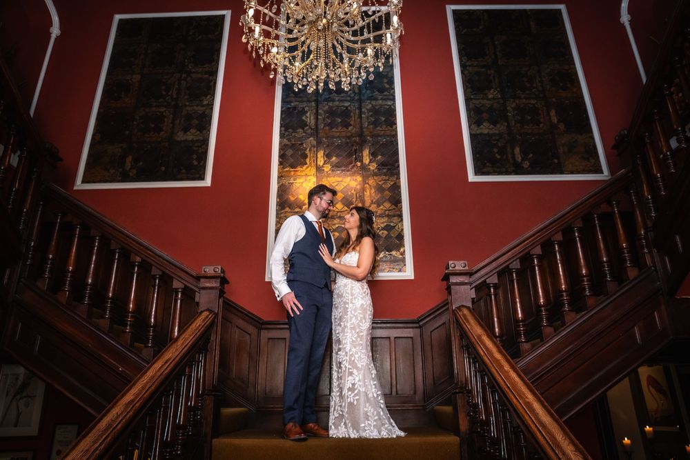Bride and groom share romantic moment on grand staircase beneath chandelier in elegant red-walled wedding venue.