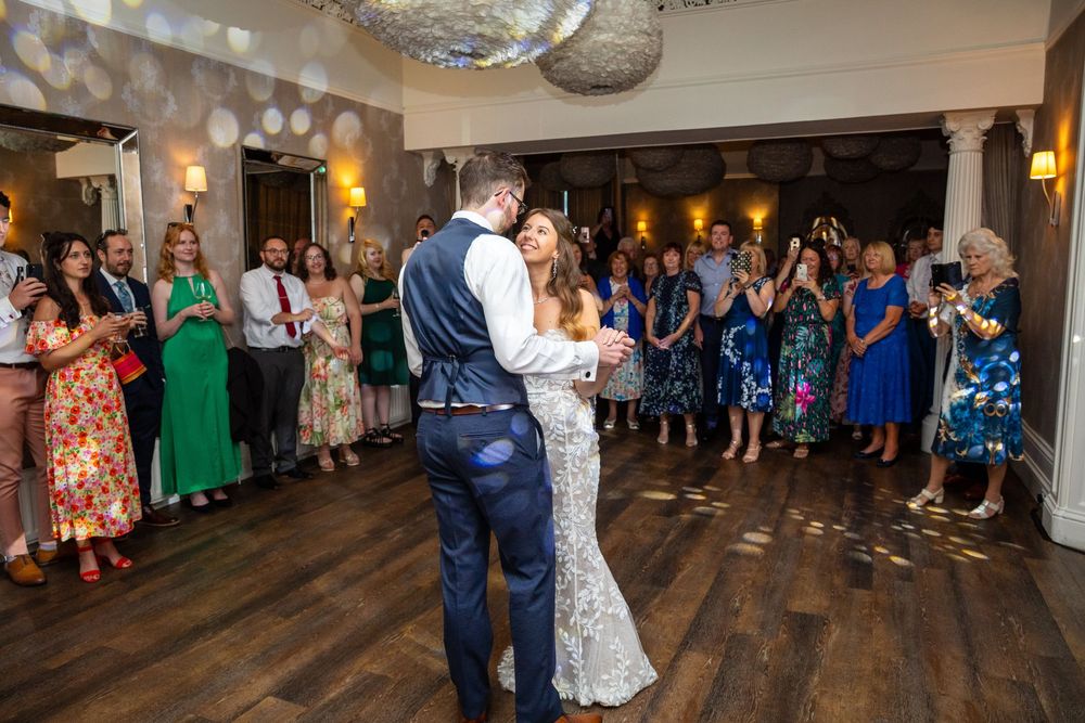 Bride and groom share first dance surrounded by cheering wedding guests in decorated reception hall.