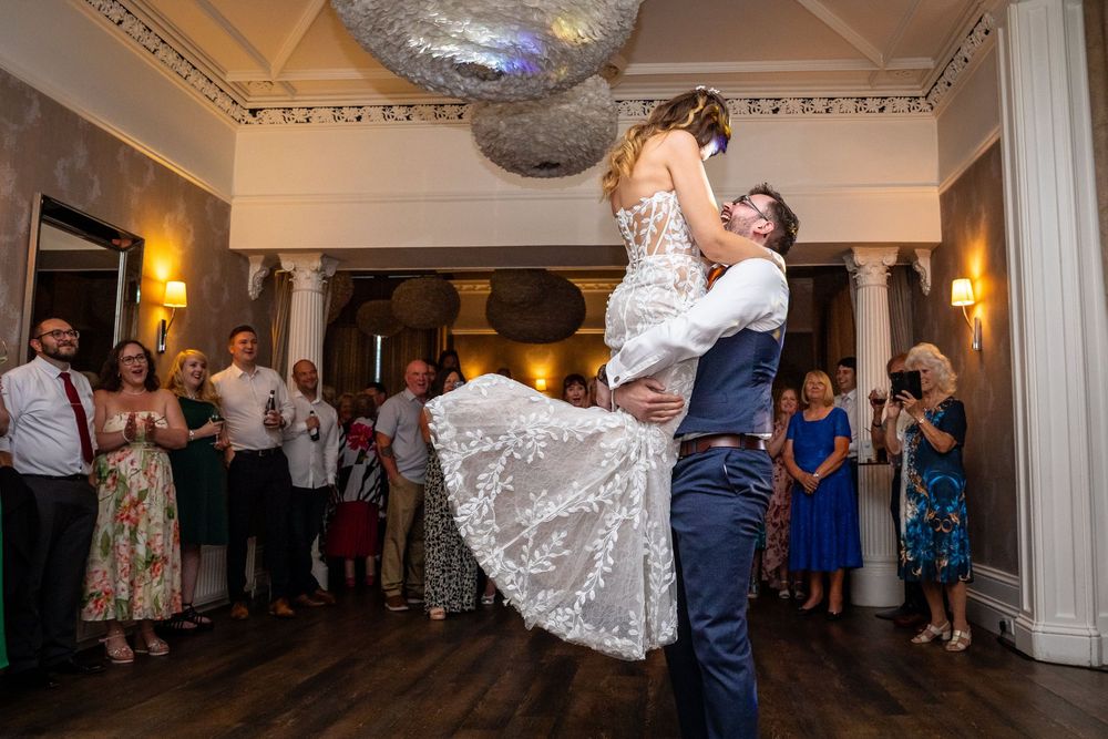 Groom lifts bride joyfully during first dance as wedding guests celebrate in elegant reception venue.