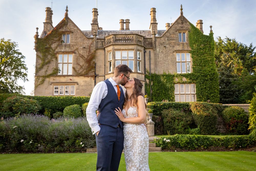Bride and groom share a kiss on manicured lawn in front of stunning ivy-covered English manor house.