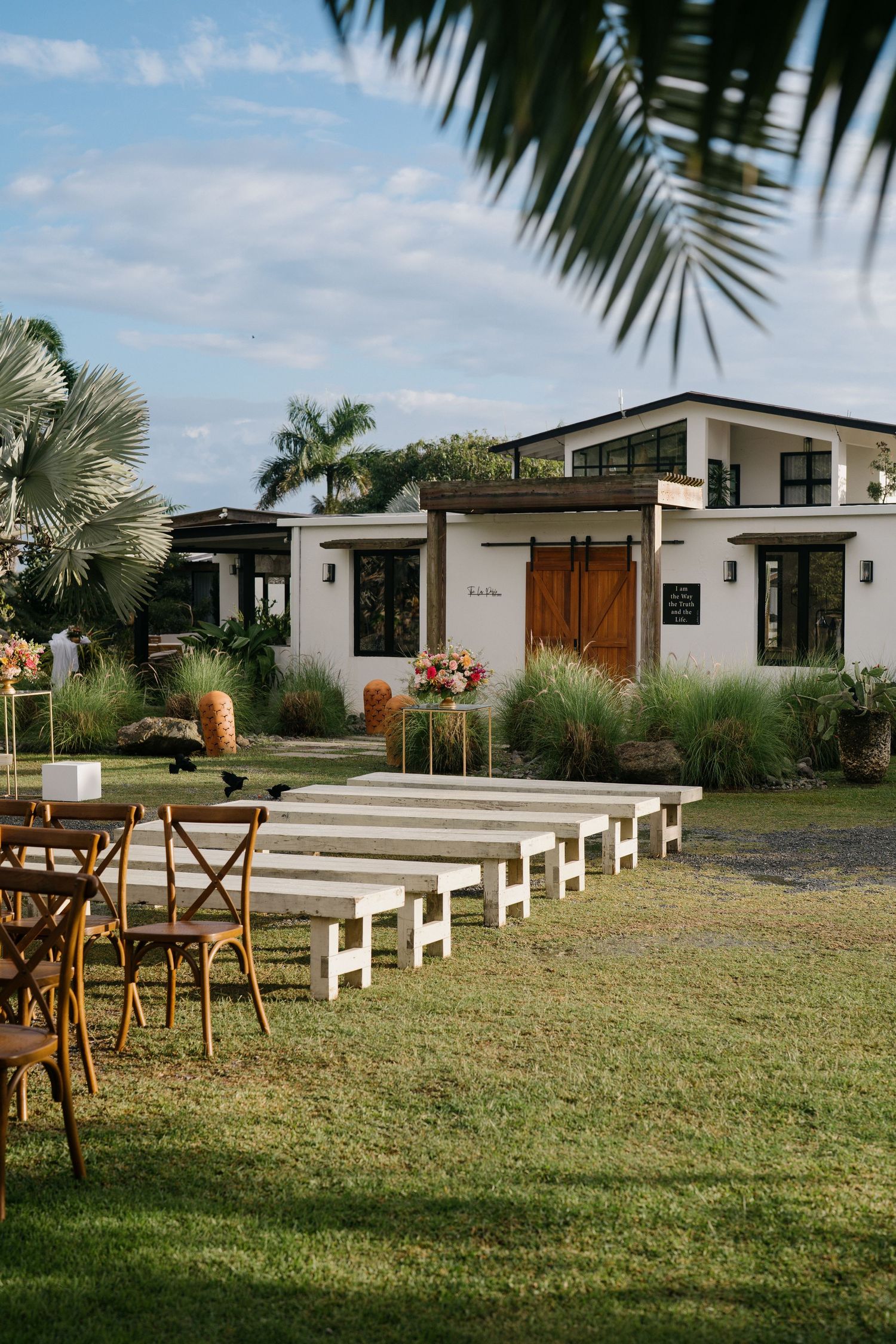Modern white tropical house with wooden accents and outdoor seating arranged on a lawn with palm trees in the background.
