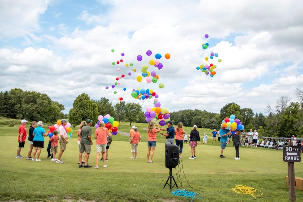 Colorful balloons float into the sky as a group gathers in a green field for a celebratory release event.