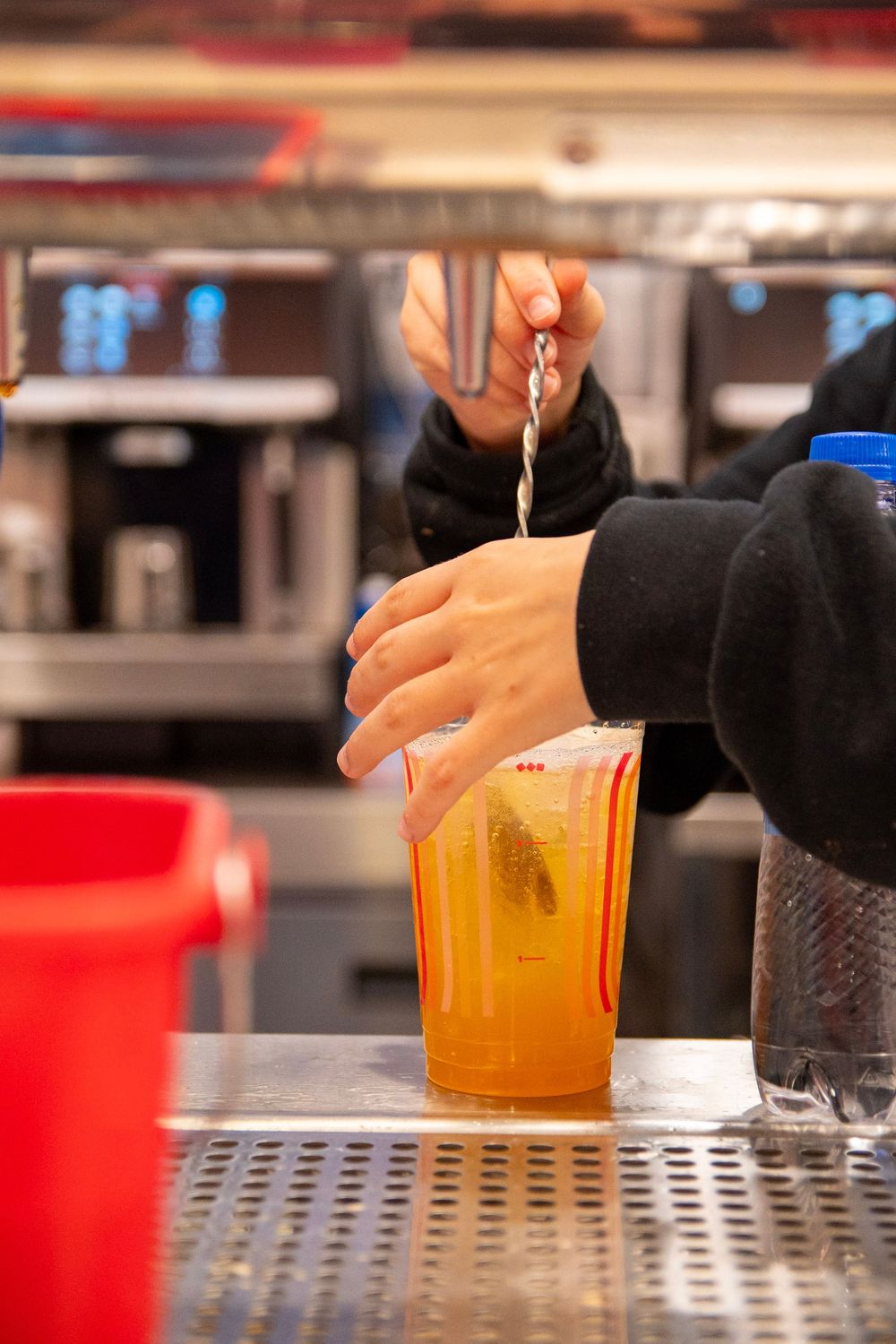 Bartender stirring an orange cocktail drink in a plastic cup on a metal bar counter.