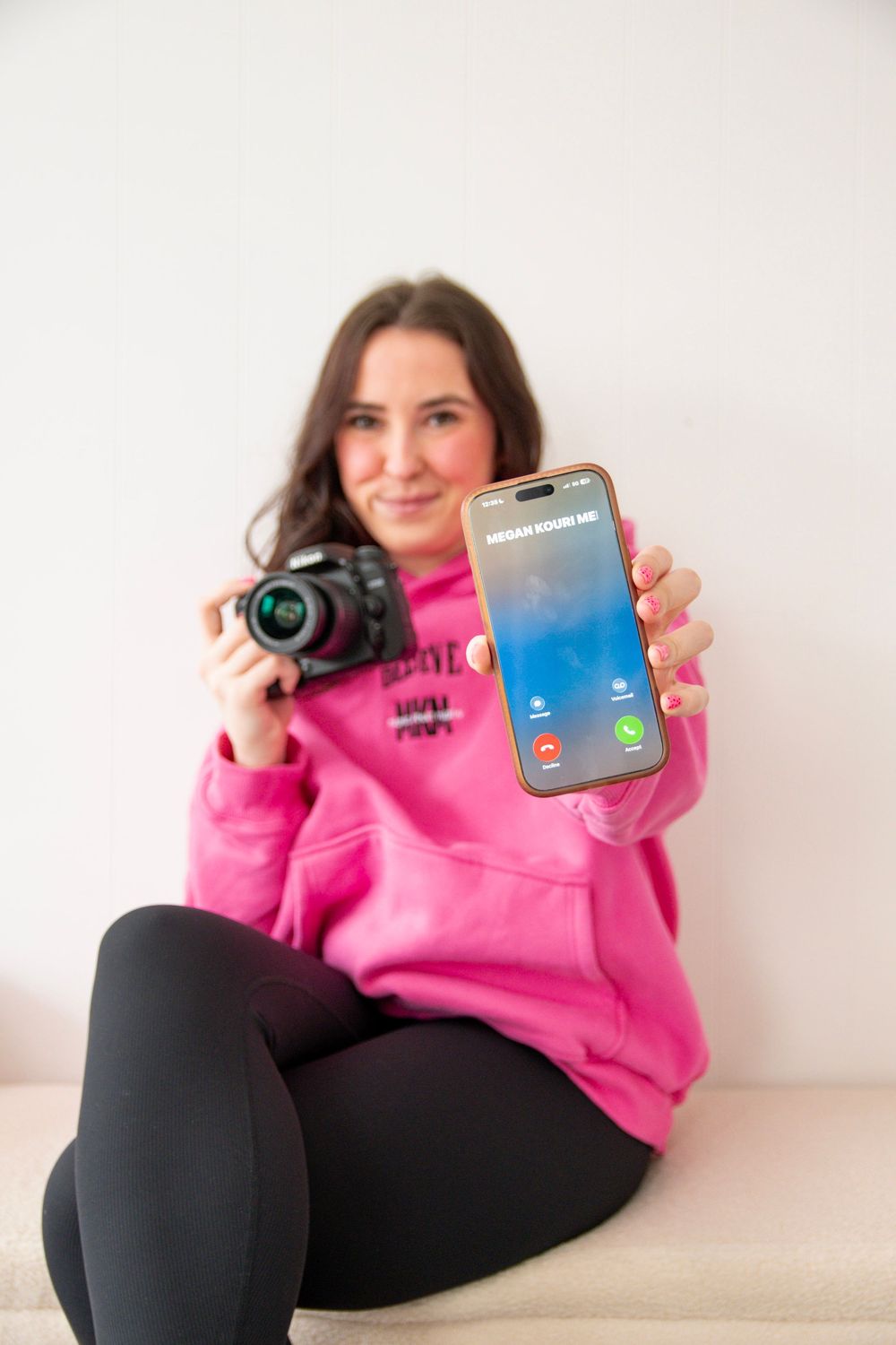 Person in pink hoodie and black pants relaxing on couch while holding camera and displaying mobile device.