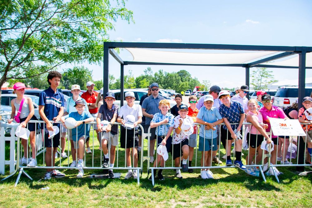 A large crowd of spectators lines up behind a metal barrier fence at an outdoor event on a sunny day.