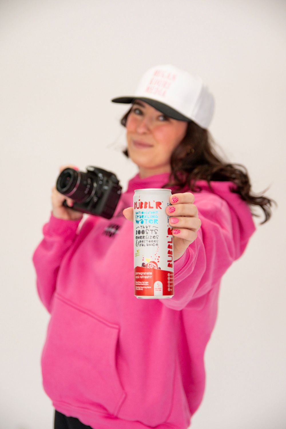 Person in bright pink sweatshirt and baseball cap holding camera and beverage can against white background.