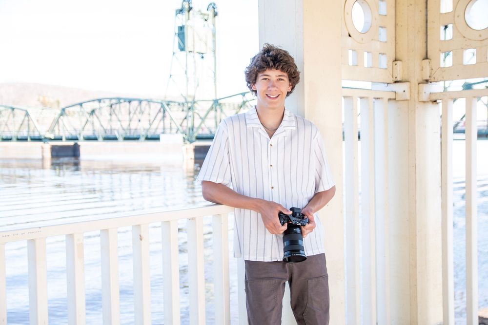 Young man smiling and holding a camera on a waterfront balcony with a green bridge in the background.