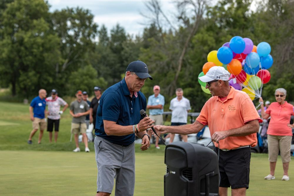 Golfers and spectators gather on a sunny golf course with colorful balloons decorating the outdoor event.