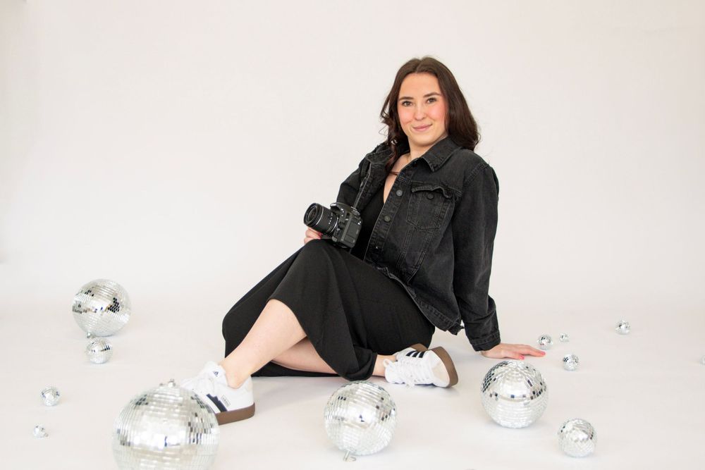 A woman in black clothing poses elegantly with silver disco balls in a minimalist white studio setting.