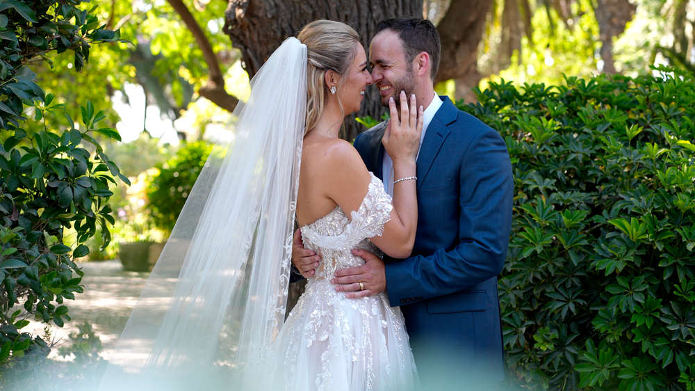 A couple shares an intimate moment in a lush garden setting with sunlight filtering through the trees.