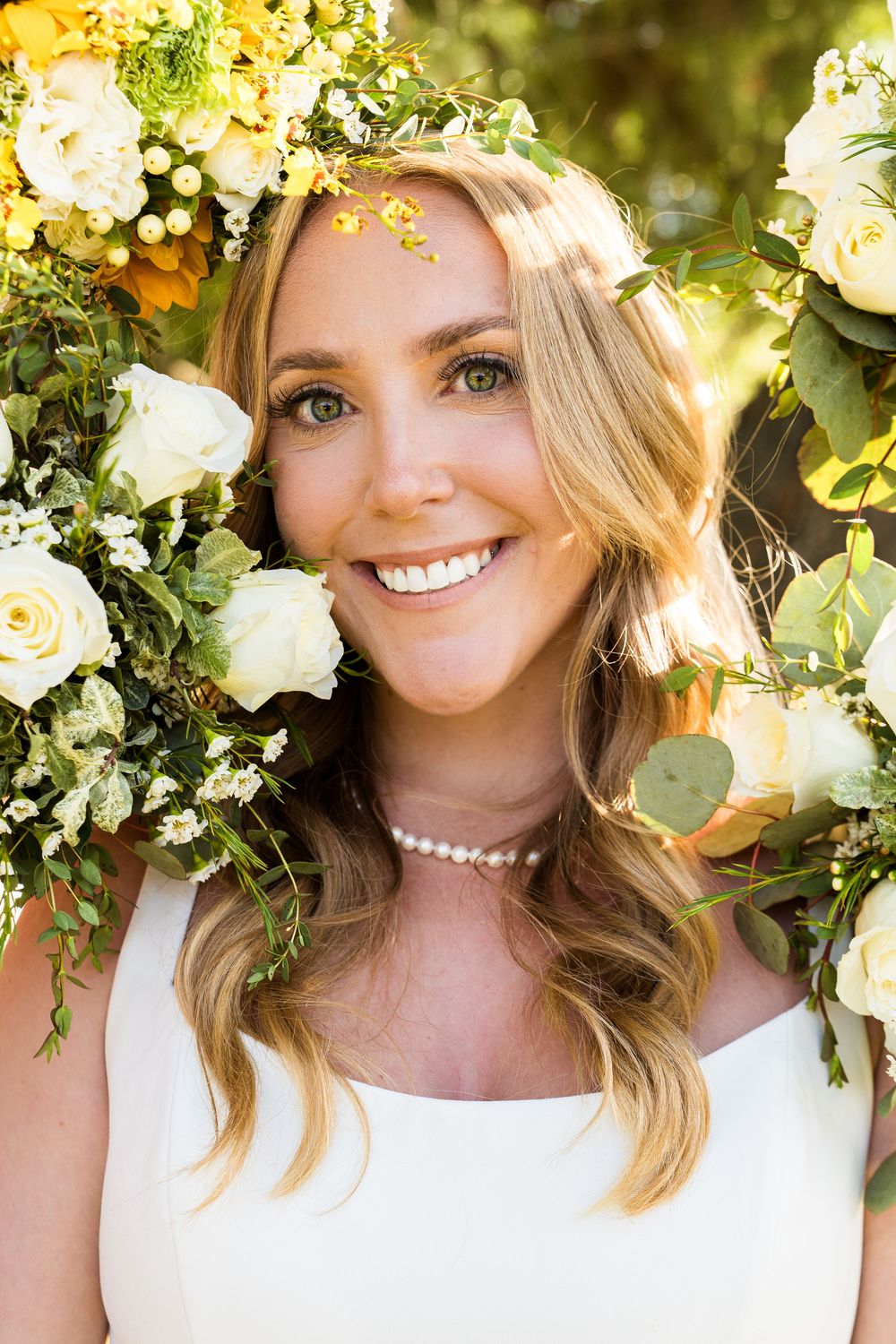A close-up of a bride wearing a floral crown and pearl necklace against a backdrop of white roses and greenery.