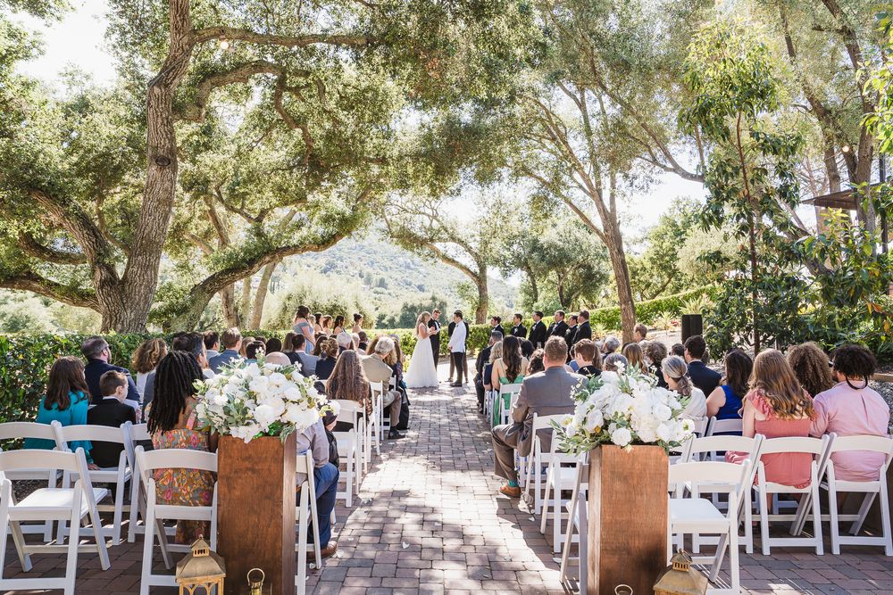 Panoramic view of an outdoor wedding ceremony setup with white chairs under shady trees on a sunny day.
