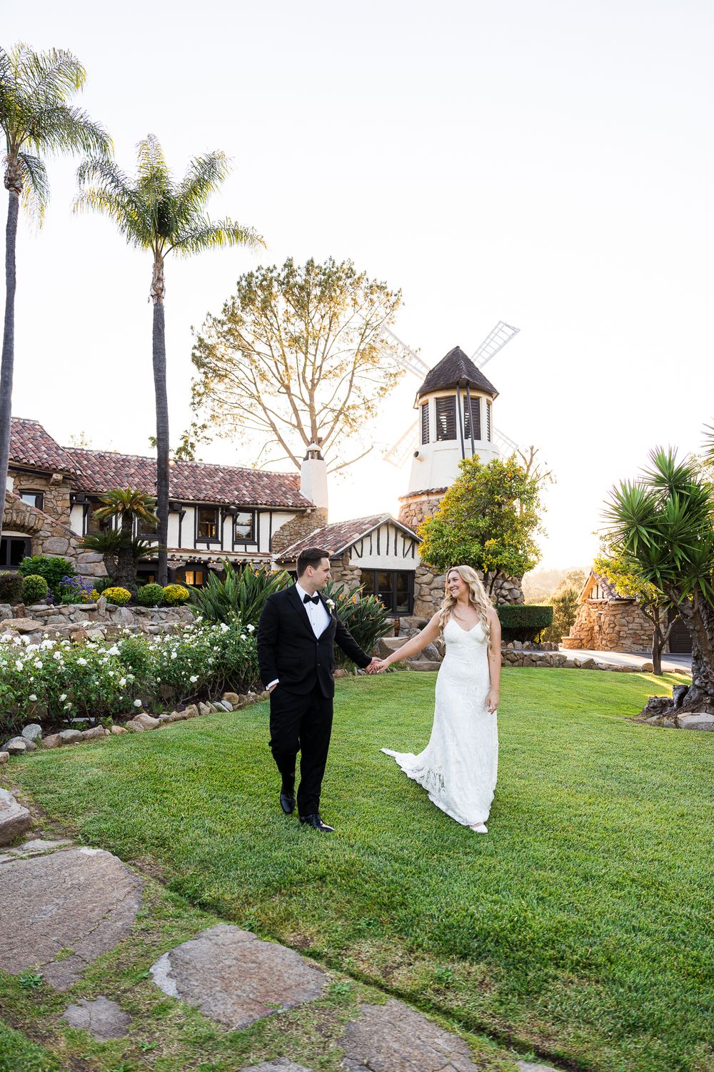A couple in formal wedding attire playfully runs and spins on a grassy lawn with palm trees and buildings in the background.