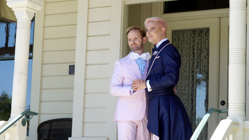 People gathered on a white porch of a house, with some wearing formal attire including a navy suit and pink dress.