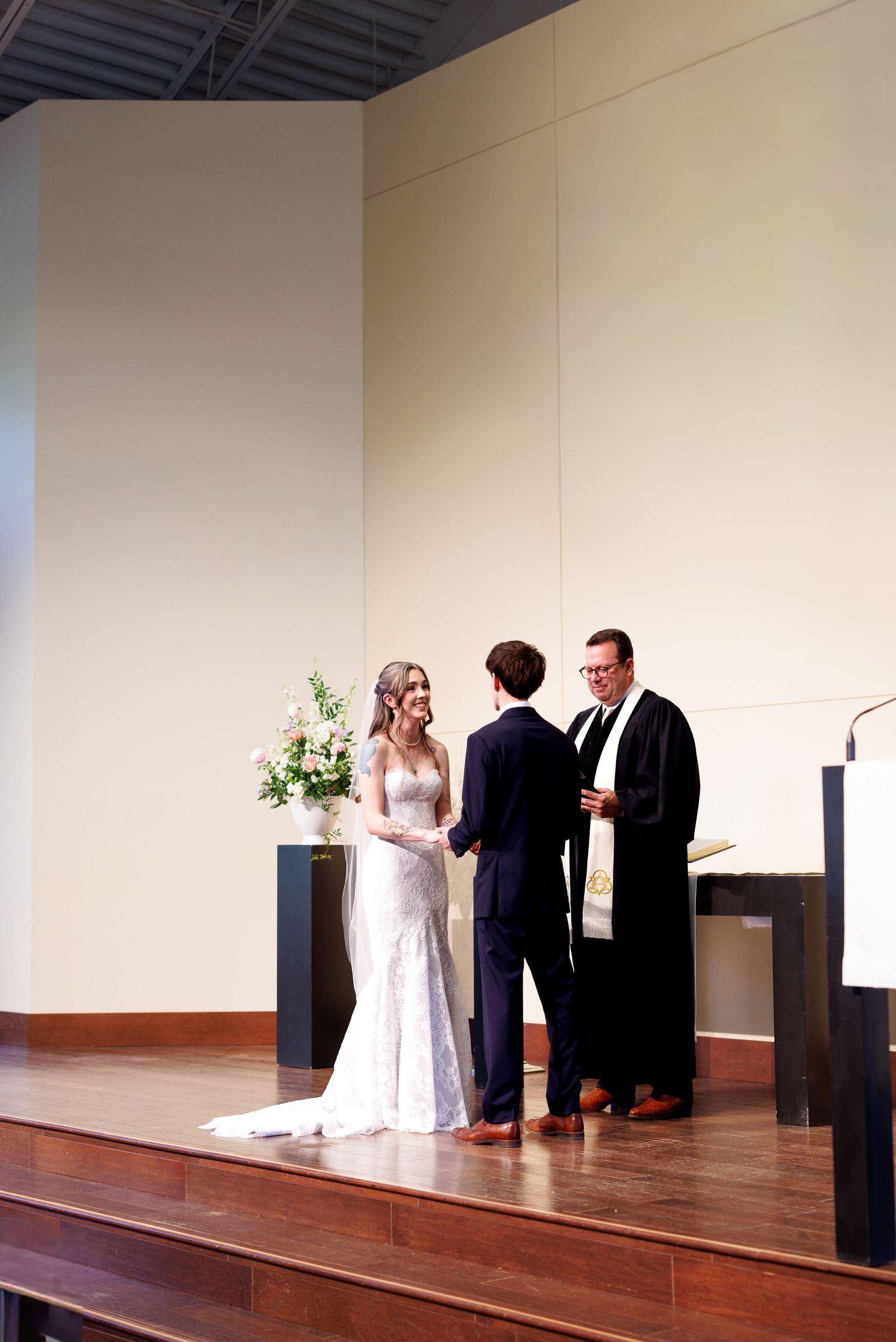 A wedding ceremony takes place on a wooden stage with minimalist decor and a floral arrangement in a modern venue.