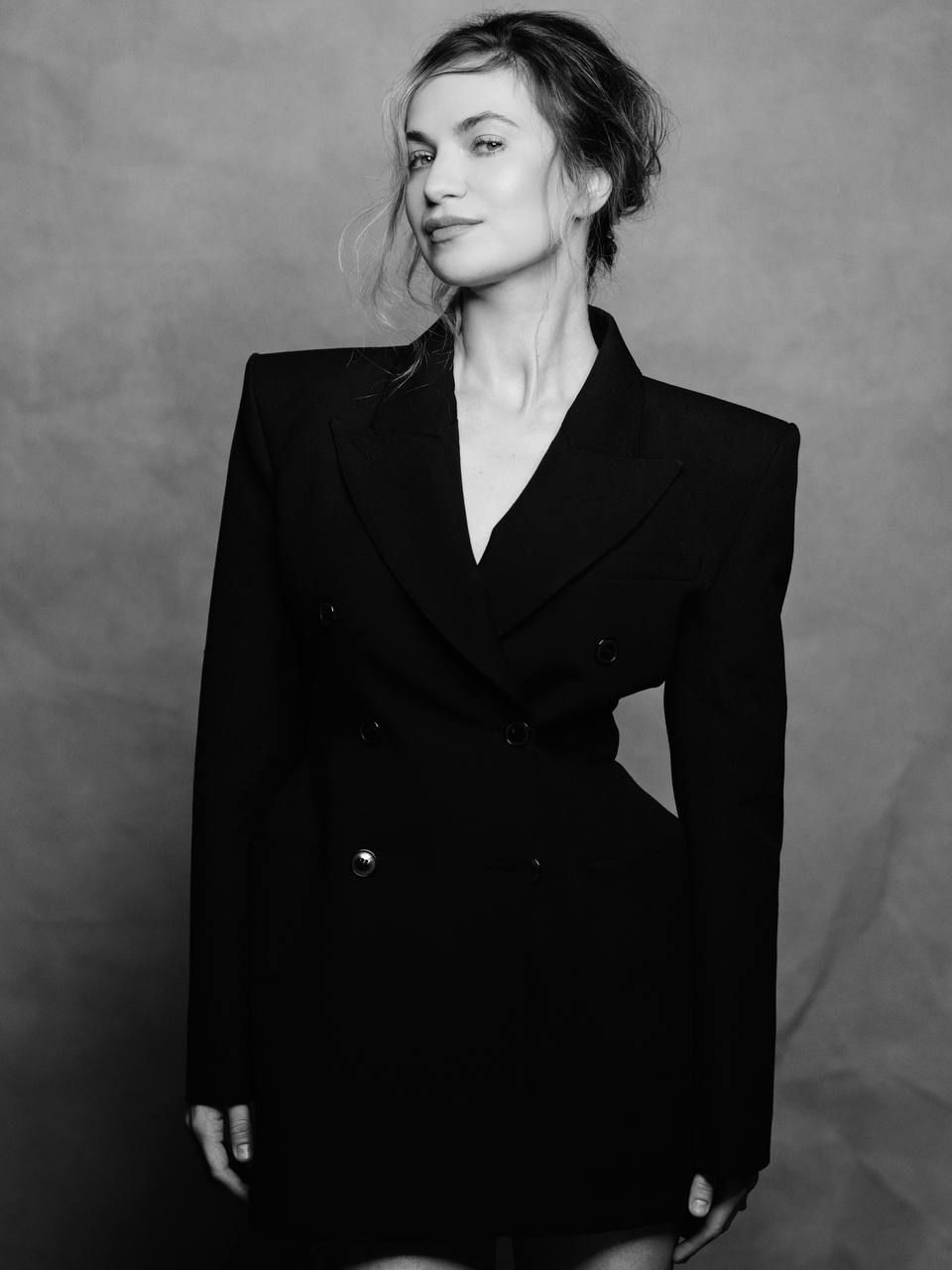 Professional photographer in beige trench coat and black dress holds camera while posing against light stone wall.
