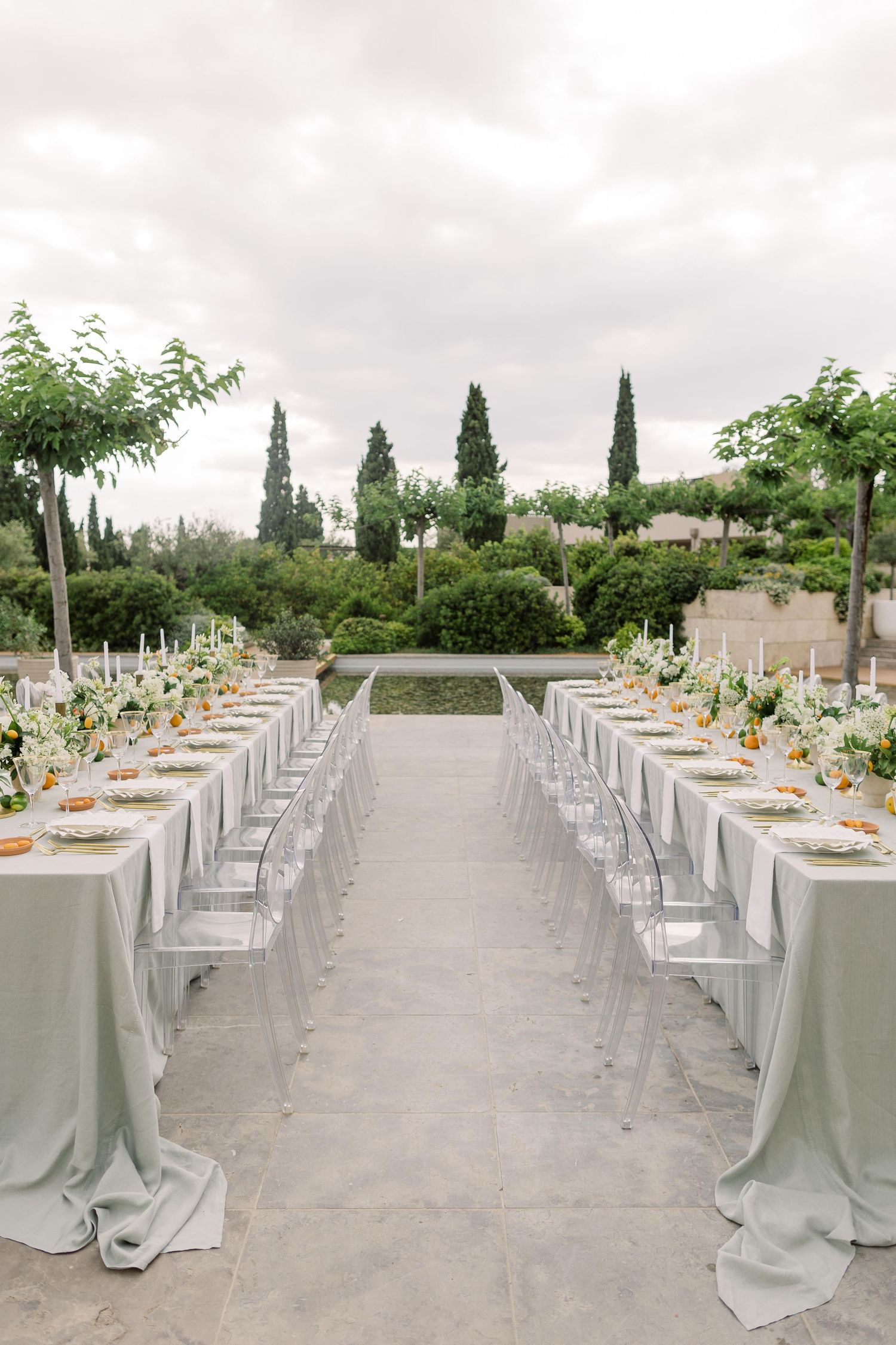 Elegant outdoor wedding reception setup with long tables covered in light gray linens and ghost chairs in a garden setting.