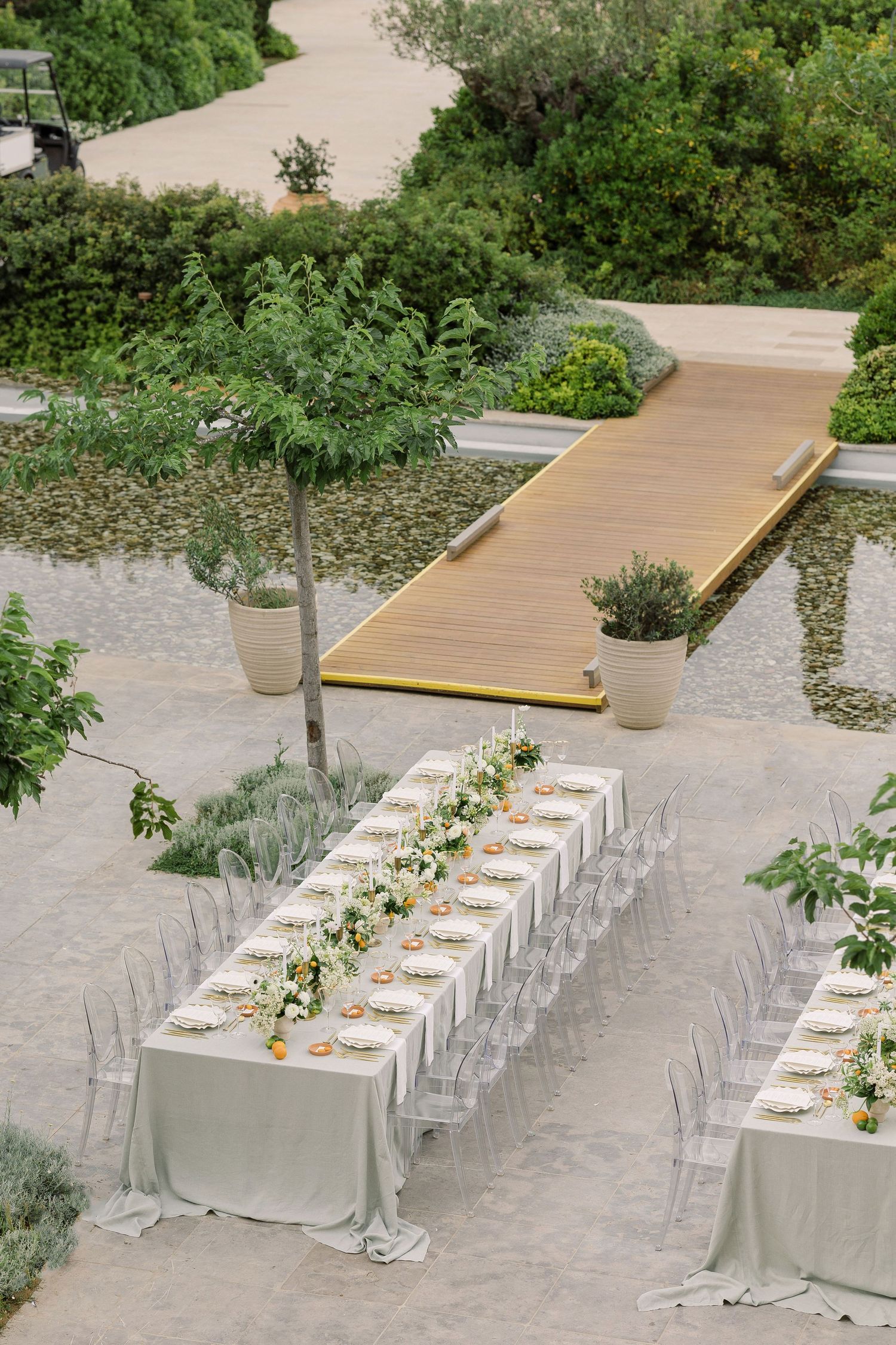 Elegant outdoor wedding reception setup with long tables, ghost chairs, and modern landscaping in a garden setting.