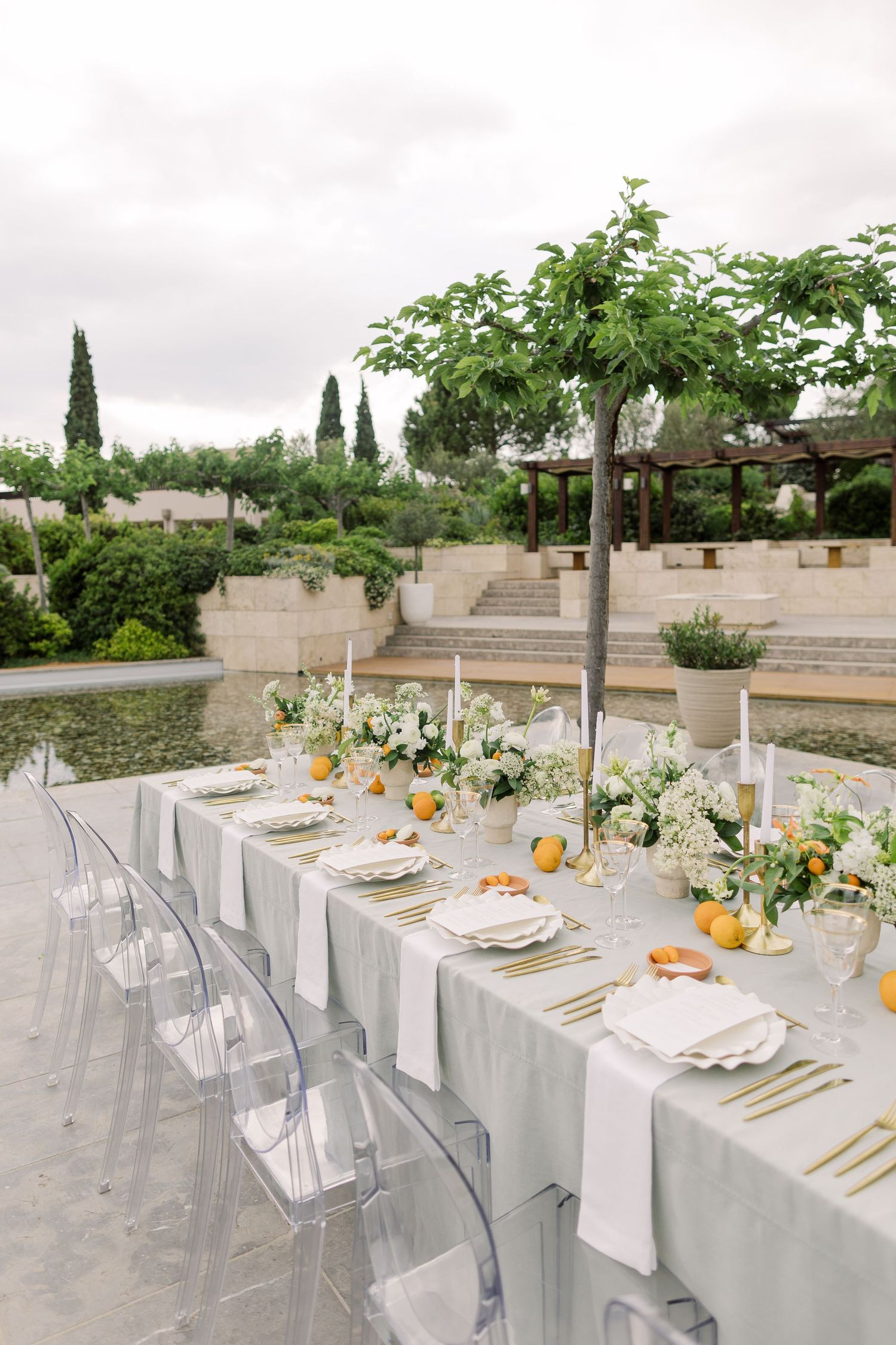Elegant outdoor wedding reception table with white linens, ghost chairs, and citrus accents set against a garden backdrop.