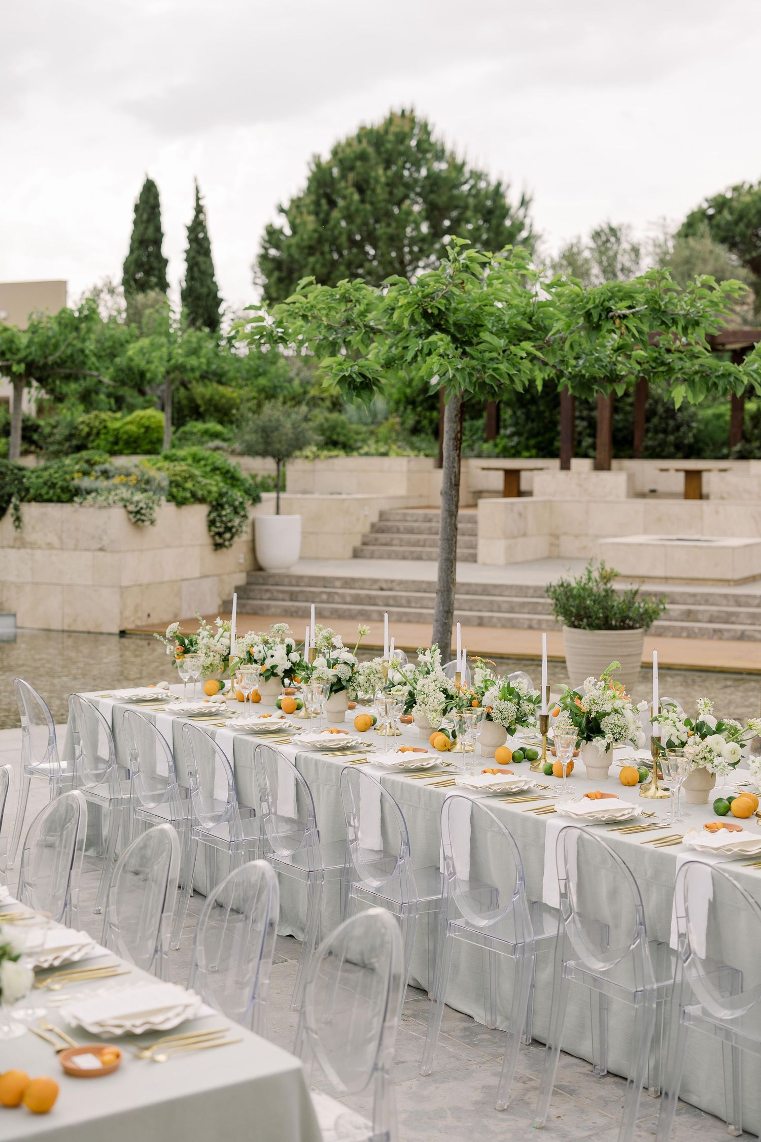 Modern garden wedding reception with clear chairs and long tables set against terraced stone steps and greenery.