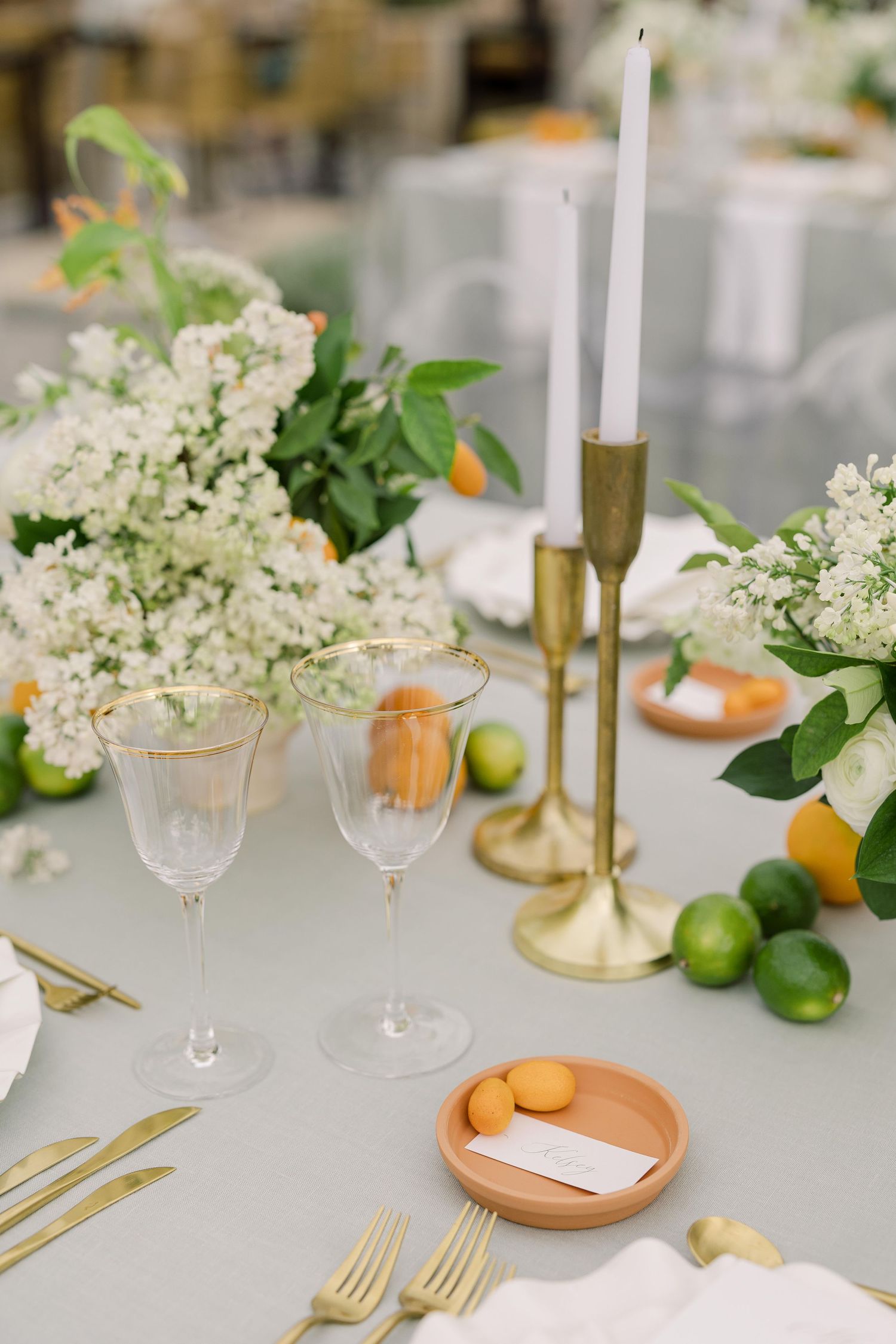 Elegant table setting with white flowers, citrus fruits, gold candlesticks and crystal glassware on gray linens.