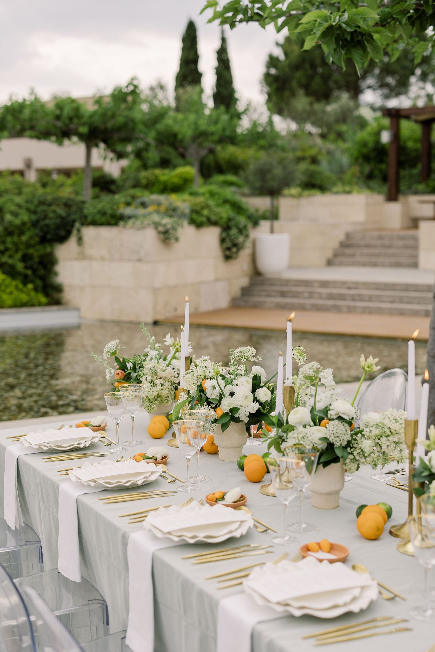 An elegant outdoor table setting with white linens, gold flatware, and fresh citrus accents on a stone patio.