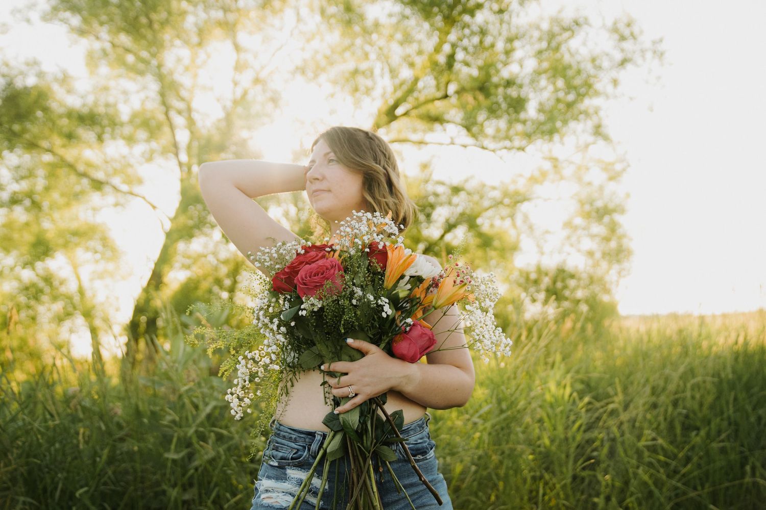 Young woman holding colorful bouquet of roses in sunny green field, golden hour light filtering through trees.
