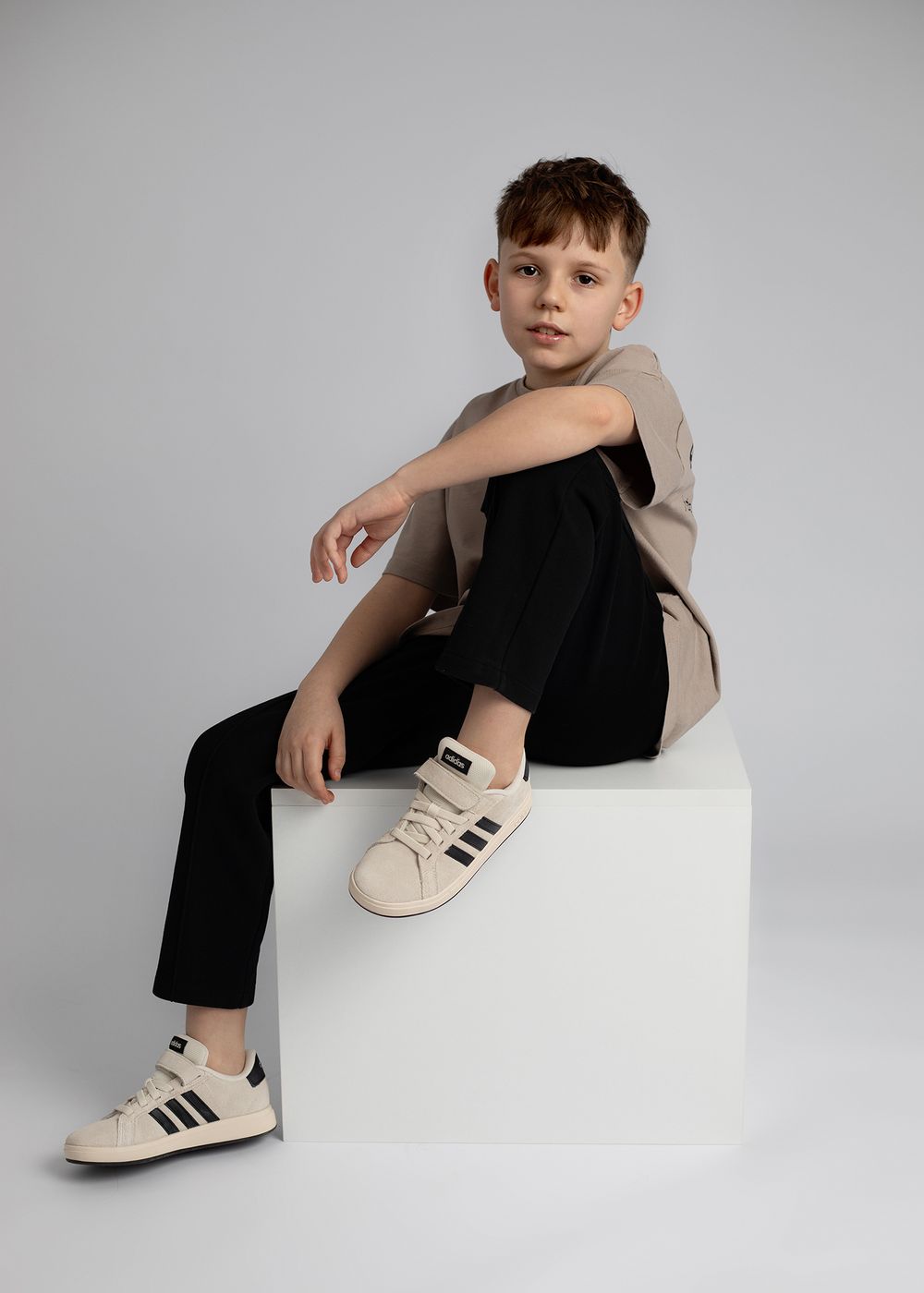 Child model wearing beige jacket and black pants poses on white cube wearing cream and black sneakers.