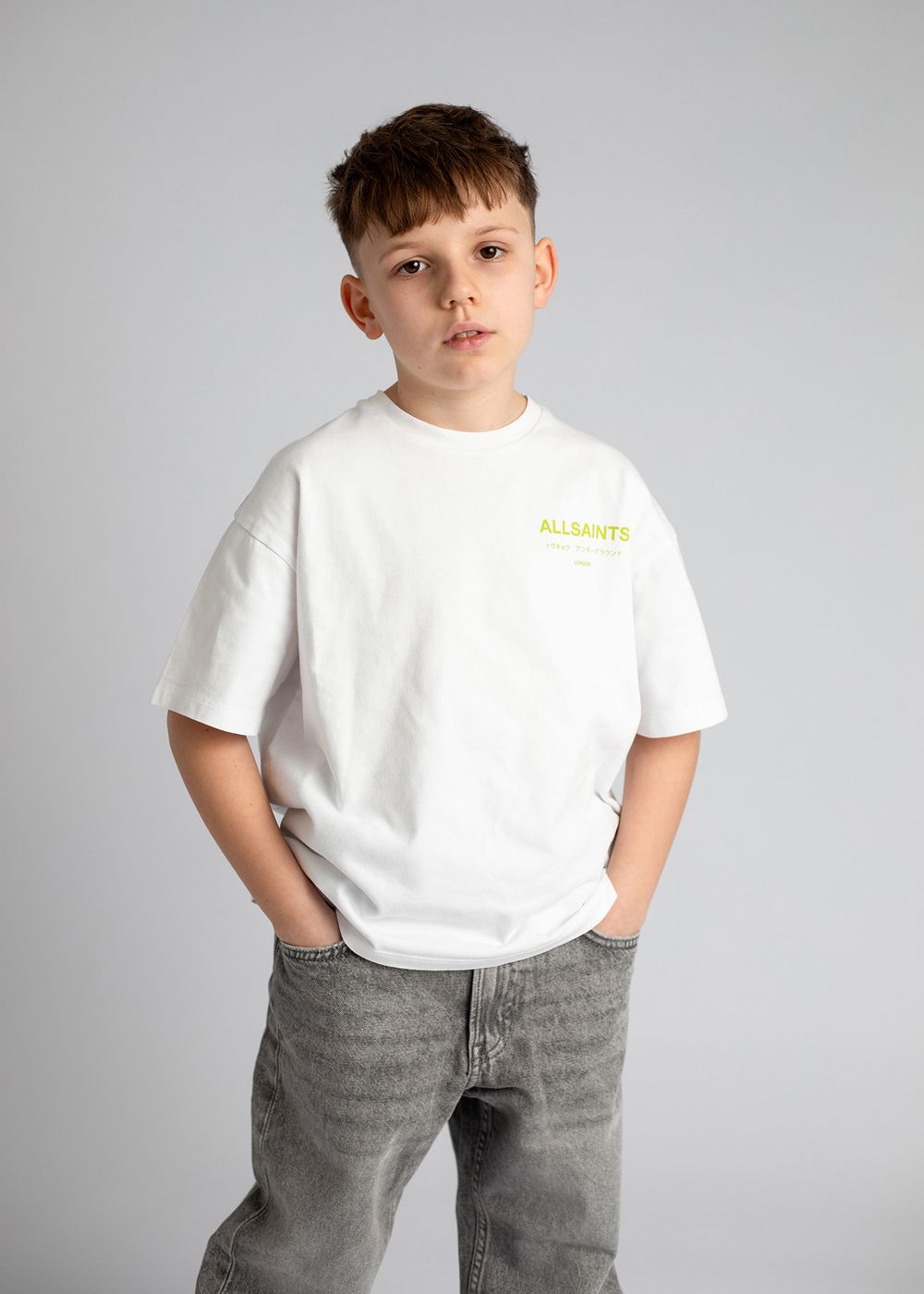 A young person wearing a white t-shirt and gray jeans poses against a light gray studio background.