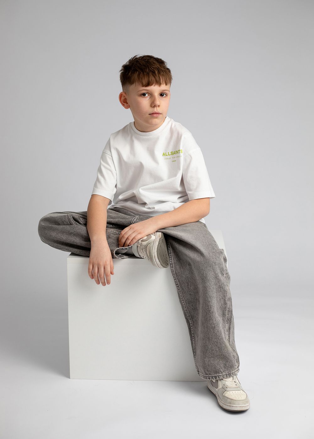 Young person in white t-shirt and gray pants sits casually on white cube pedestal against light gray studio background.