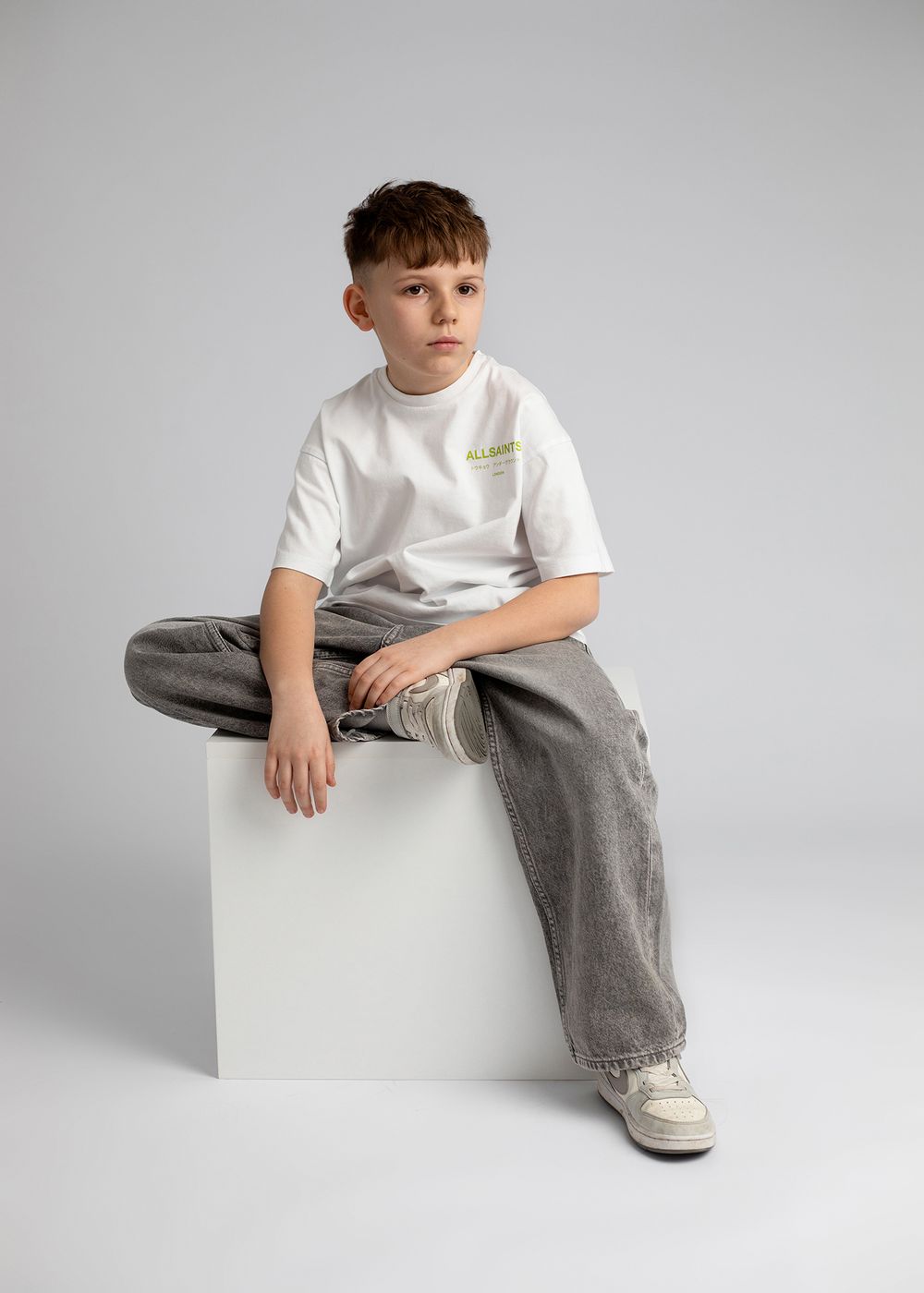 A person in a white t-shirt and gray pants sits casually on a white cube against a light gray studio backdrop.