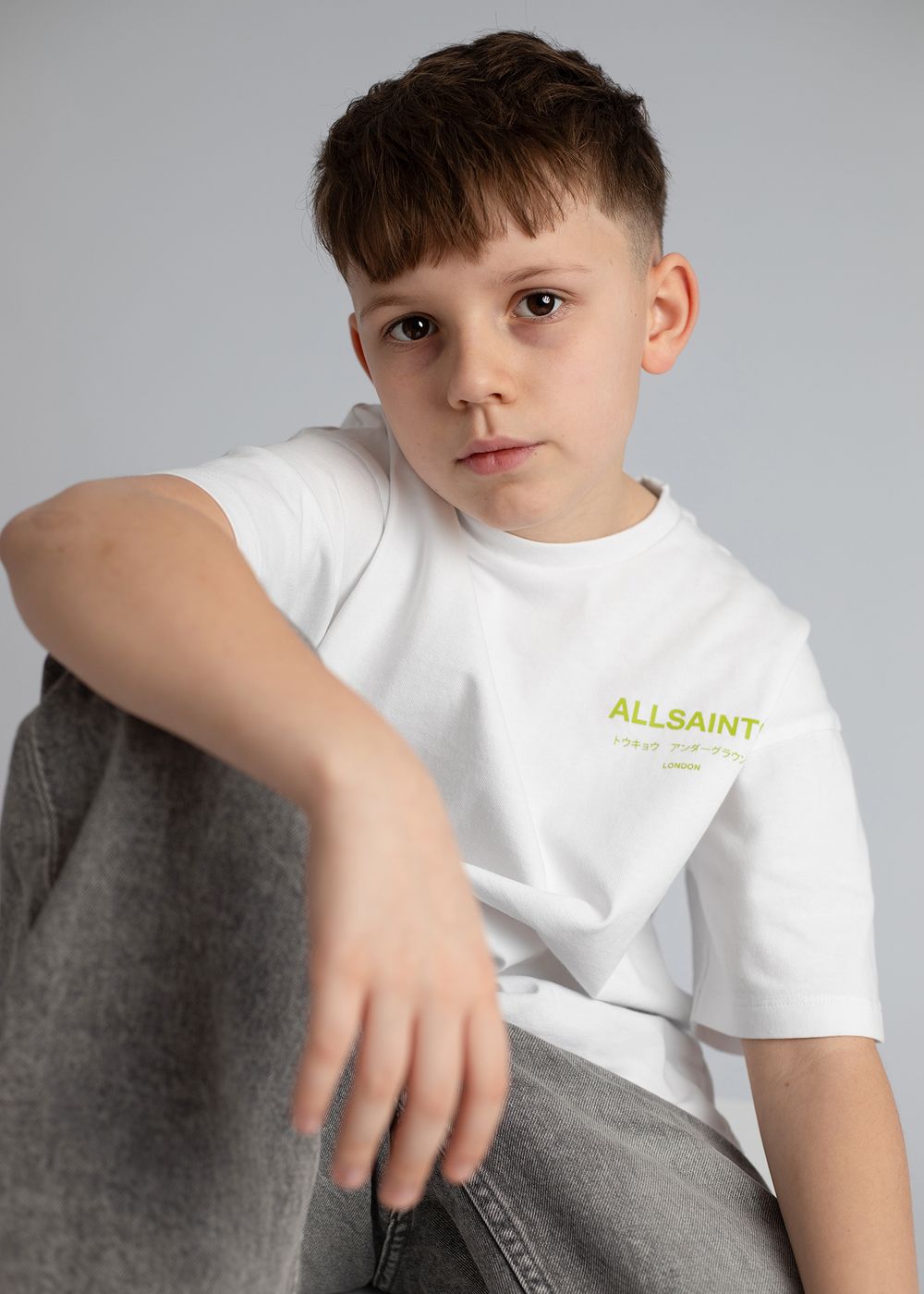 A young child in a white t-shirt sits casually on a gray couch in a minimalist setting.