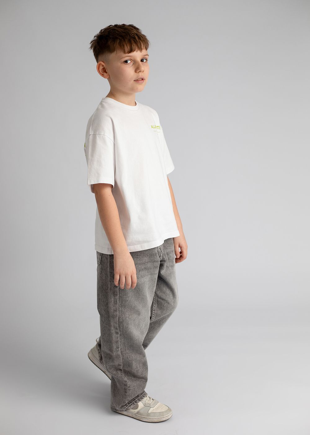 Young child wearing a white t-shirt and gray pants poses in various angles against a light gray studio background.