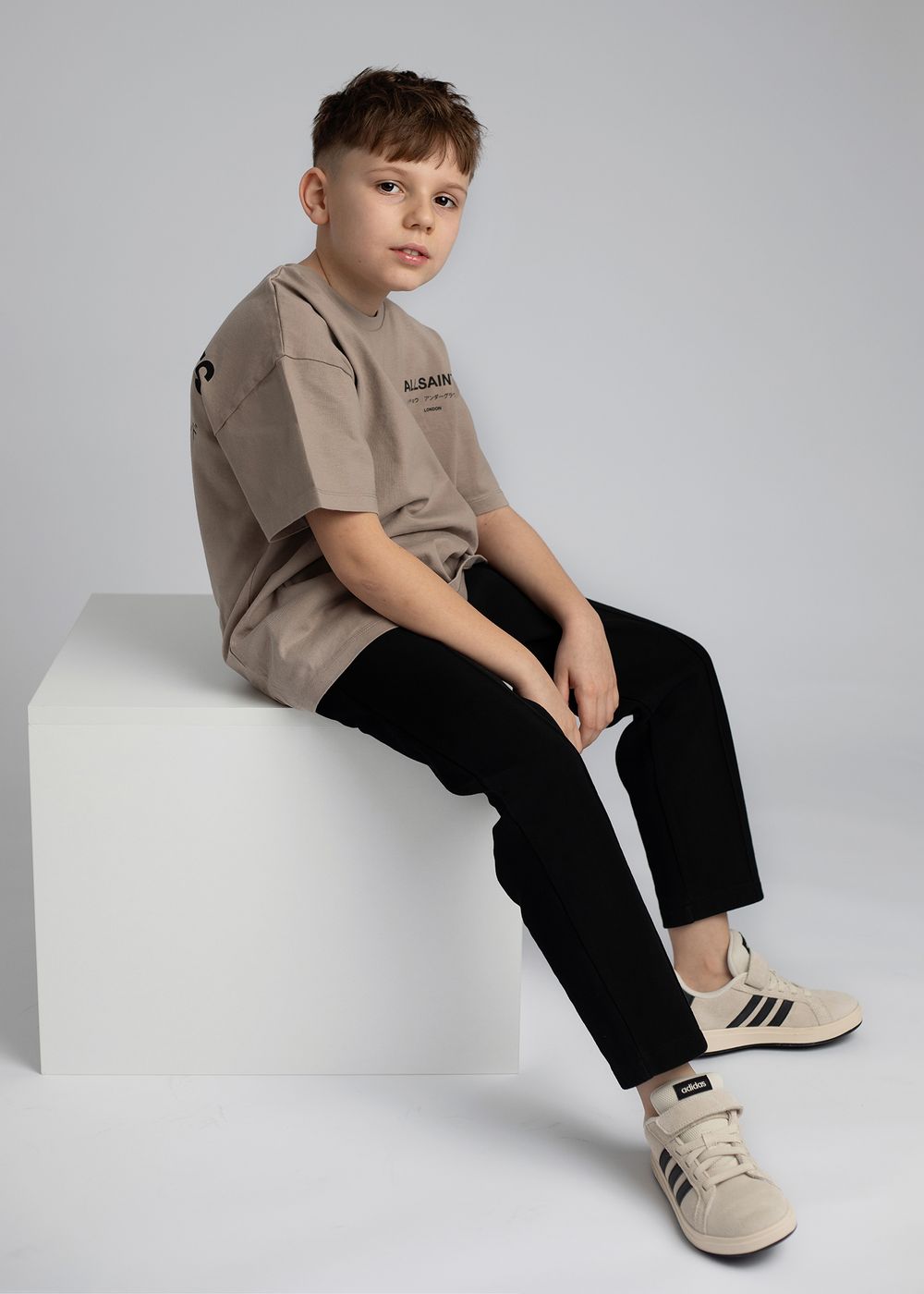 Young child in beige shirt and black pants sits casually on white surface wearing cream-colored sneakers.