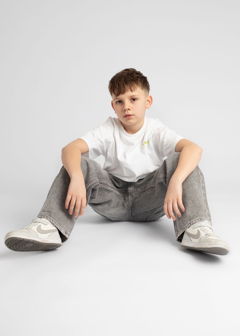 A person in a white shirt and gray pants sits on the floor in a relaxed pose against a white background.
