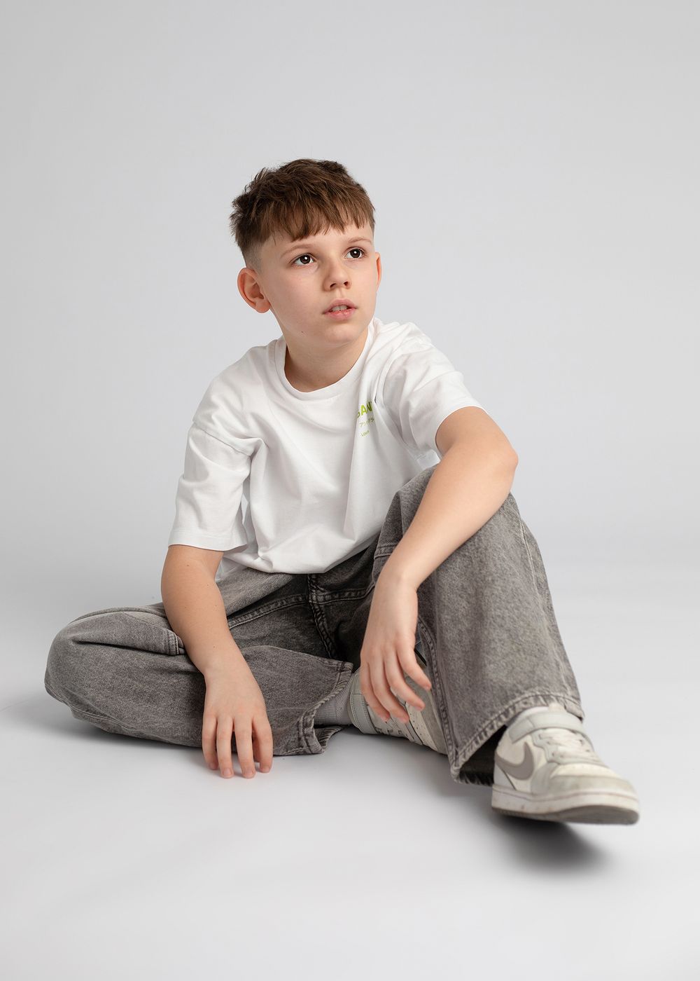 Child in white t-shirt and gray pants lounges on floor in casual studio portraits against light background.