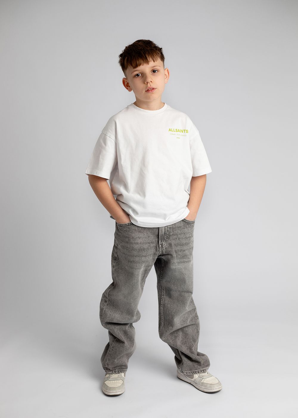 Young person wearing a white t-shirt and baggy gray jeans poses against a light gray studio backdrop.
